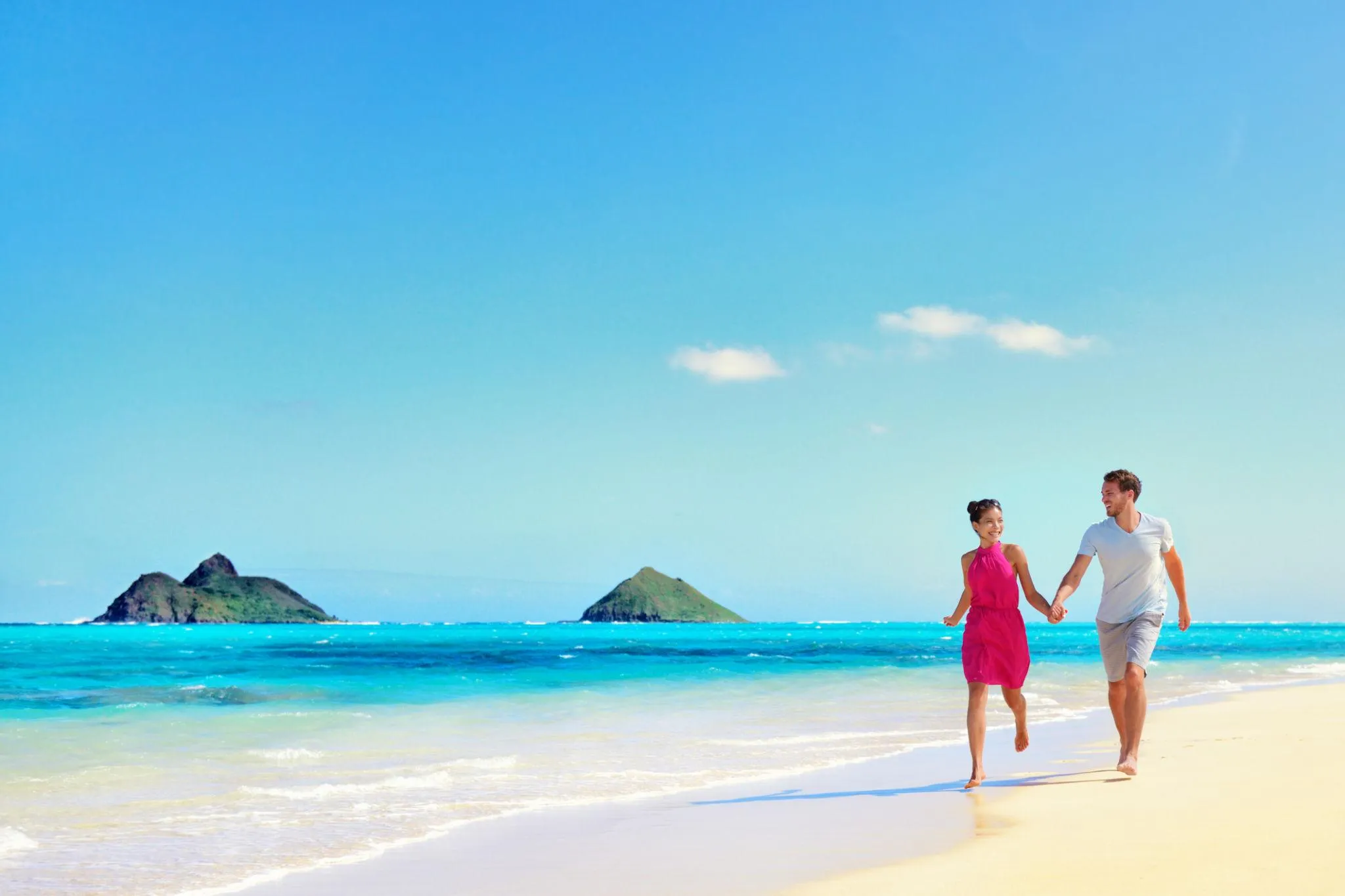A couple walks along a sandy Oahu beach with the Mokulua islets offshore