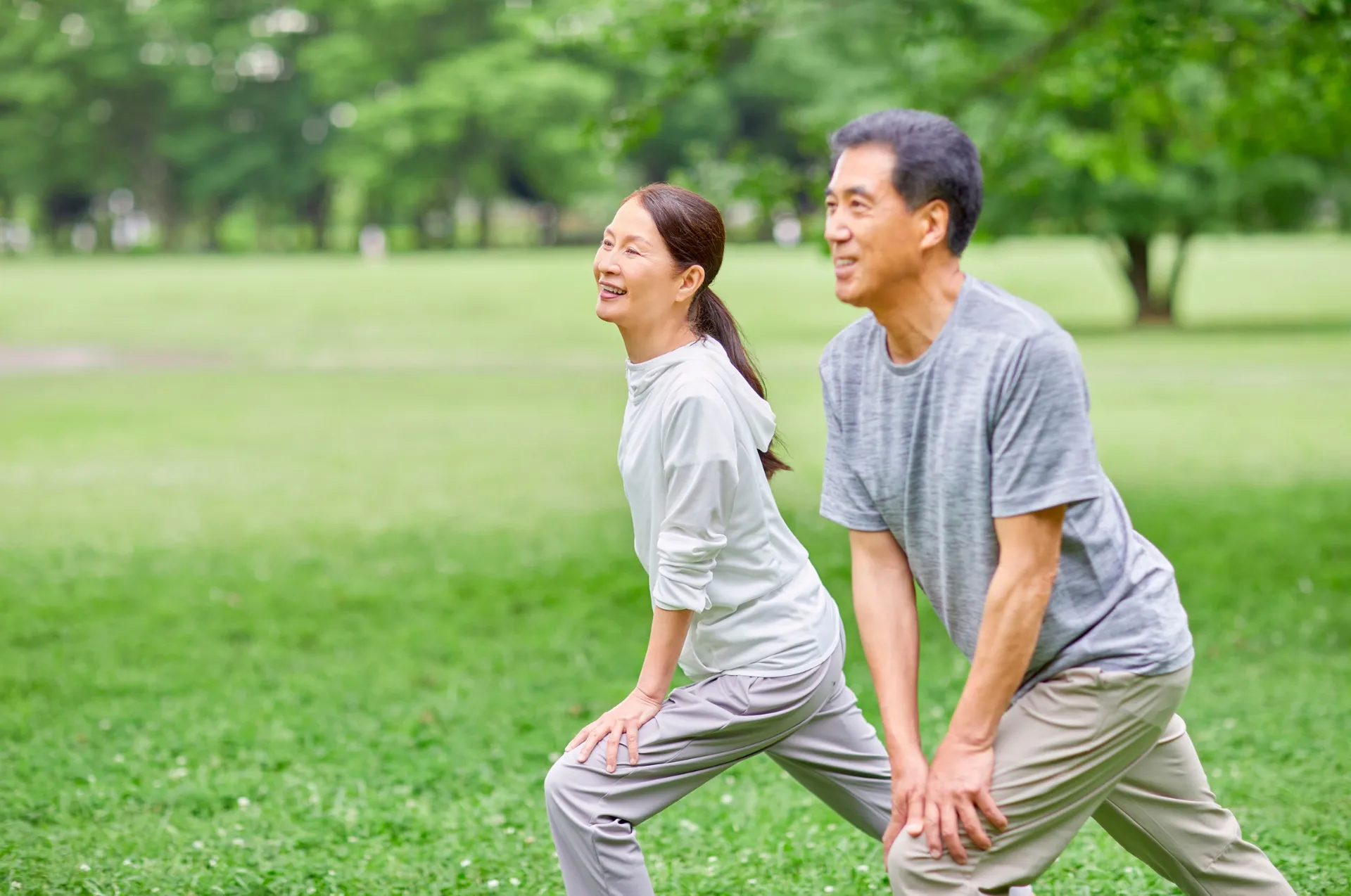 An older couple does lunging stretches together in a grassy park